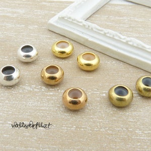 Könnte beinhalten: Sortiment von acht runden Metallperlen in verschiedenen Farben, darunter Gold, Silber und Messing. Die Perlen haben eine glatte, glänzende Oberfläche und sind auf einer hellbraunen Oberfläche angeordnet.