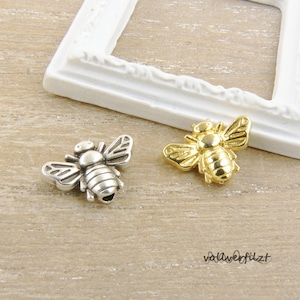 Könnte beinhalten: Zwei Bienen-Anhänger, einer silber und einer gold, mit detaillierten Flügeln und Körpern. Die Anhänger liegen auf einer Holzoberfläche.