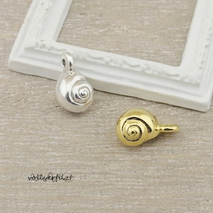 Könnte beinhalten: Zwei kleine muschelförmige Anhänger, einer silberfarben und einer goldfarben, mit einer Öse zum Anbringen an Schmuck oder andere Bastelarbeiten.