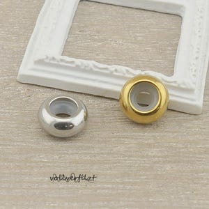 Könnte beinhalten: Zwei runde Perlen, eine silberne und eine goldene, mit weißem Innenraum. Die Perlen befinden sich neben einem weißen Bilderrahmen. Der Text "vollverfilzt" befindet sich am unteren Bildrand.