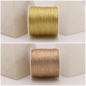 Könnte beinhalten: Zwei Spulen Metallfaden. Eine ist goldfarben, die andere bronzefarben. Der Faden ist eng auf weißen Spulen aufgewickelt. Der Hintergrund ist neutral gefärbt.