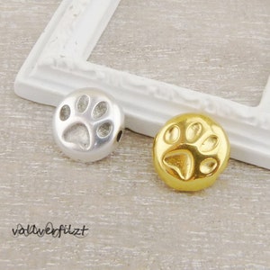 Könnte beinhalten: Zwei Metall-Pfotenabdruck-Anhänger, einer silber und einer gold. Die Anhänger sind rund und haben ein erhabenes Pfotenabdruck-Design.