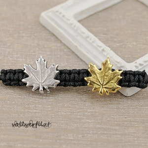 Könnte beinhalten: Ein schwarzes Kordelarmband mit zwei silbernen und goldenen Ahornblatt-Anhängern. Das Armband liegt auf einer Holzoberfläche.