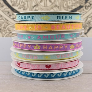 Könnte beinhalten: Ein Stapel von acht bunten Bandspulen mit verschiedenen Sprüchen darauf gedruckt, darunter "Carpe Diem", "Das Leben ist schön", "Amour", "Happy", "Good Vibes", "Love Sister" und ein Wellenmuster.