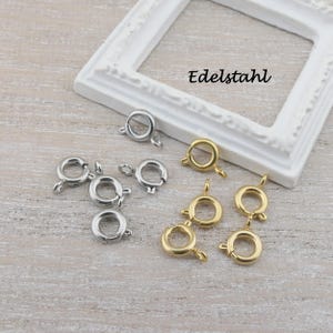Könnte beinhalten: Eine Sammlung von silber- und goldfarbenen Federringverschlüssen. Die Verschlüsse sind rund mit einem kleinen Hebel zum Öffnen und Schließen. Das Wort "Edelstahl" steht in schwarzer Schrift auf einem weißen Zierrahmen.