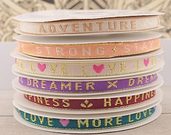 Könnte beinhalten: Ein Stapel von sechs Rollen bunten Bandes mit goldener Einfassung. Jedes Band hat eine andere Botschaft, darunter "Adventure", "Strong Stay", "Love Love", "Dreamer", "Happiness" und "Love More Love".