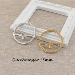 Könnte beinhalten: Zwei runde Anhänger, einer silberfarben und einer goldfarben, jeweils mit einer Vogelsilhouette in einem Ring. Der silberne Anhänger befindet sich links, der goldene rechts. Der Text "Durchmesser 15mm" steht unten.