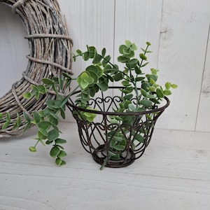 Könnte beinhalten: Ein schwarzer Metalldraht-Pflanztopf mit einem dekorativen Herzdesign. Der Topf ist mit grünen Blattpflanzen gefüllt.