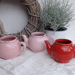 Könnte beinhalten: Drei Keramik-Teekannen in Rosa und Rot. Die Teekannen sind klein und haben einen rustikalen, Vintage-Look.