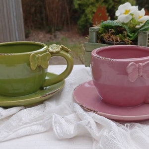 Könnte beinhalten: Zwei Keramik-Teetassen mit Untertassen, eine grün und eine rosa, verziert mit Schmetterlingsakzenten. Die Teetassen sind im Vintage-Stil mit einer abgenutzten Oberfläche.