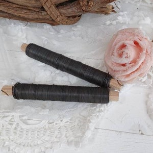 Könnte beinhalten: Zwei Spulen schwarzen Drahtes mit Holzenden, die auf einer weißen Spitzenunterlage und einer weißen Holzoberfläche liegen. Im Hintergrund befindet sich eine rosa Stoffrose.