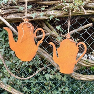 Könnte beinhalten: Zwei orangebraune Teekannen-Ausschnitte aus Metall, die an Schnüren hängen. Die Teekannen haben ein rustikales, verwittertes Aussehen und hängen vor einem Hintergrund aus Grün und einem Maschendrahtzaun. Dekorative Gartendekoration.
