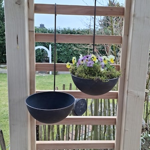 Könnte beinhalten: Zwei schwarze Hängepflanzenbehälter mit Metallketten, einer gefüllt mit violetten und gelben Stiefmütterchen und grünem Moos, der andere leer, hängen an einem Holzlatten-Gitter.
