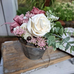 Könnte beinhalten: Ein Blumenarrangement in einem kleinen Eimer aus verzinktem Metall mit einer großen cremefarbenen Rose, kleineren weißen Rosen, rosa Füllblumen, staubgrünen Eukalyptusblättern und einer altrosa Satinschleife zwischen den Blüten.