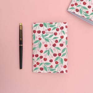 Può includere: Un quaderno rosa con una stampa di ciliegie e la parola "NOTES" sulla copertina. Una penna nera è accanto al quaderno su una superficie rosa.