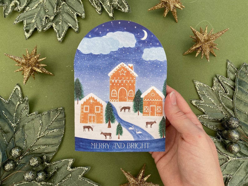 Merry and Bright snowglobe card. 3 decorated gingerbread houses in the snow. Swans in the river. Reindeers and fir trees in the on land. Clouds with snow, moon and stars above.
