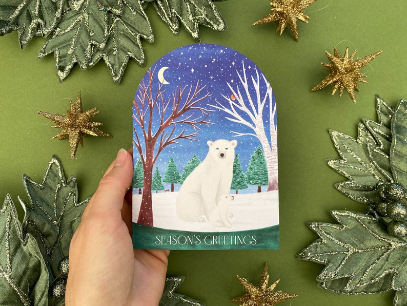 Season’s greetings snowglobe card with polar and baby cub in a woodland with a robin and squirrel in the background. Starry with a crescent moon and snow.