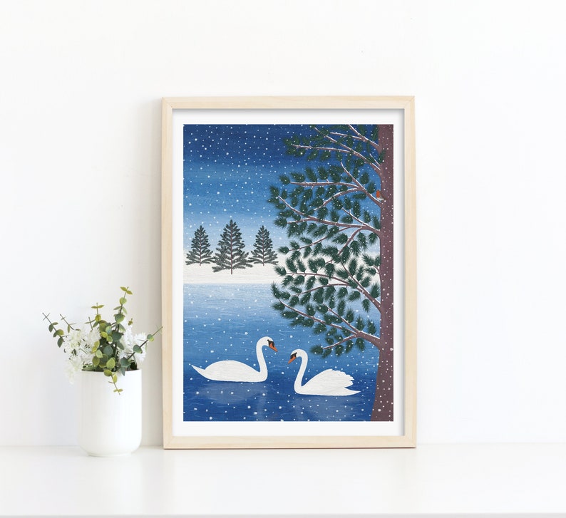 Christmas art print with mock up oak frame with 2 swans in a lake, a tree in the foreground with a robin perched on a branch and 3 christmas trees in the background. The daytime blue sky background and falling snow.