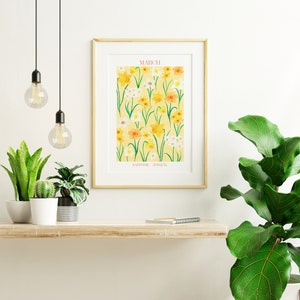 May include: A framed print with a yellow background and a pattern of yellow and white daffodils and jonquils. The print has the word "MARCH" and the words "DAFFODIL, JONQUIL" at the bottom. The print is hanging on a white wall above a wooden shelf with plants and a notebook.