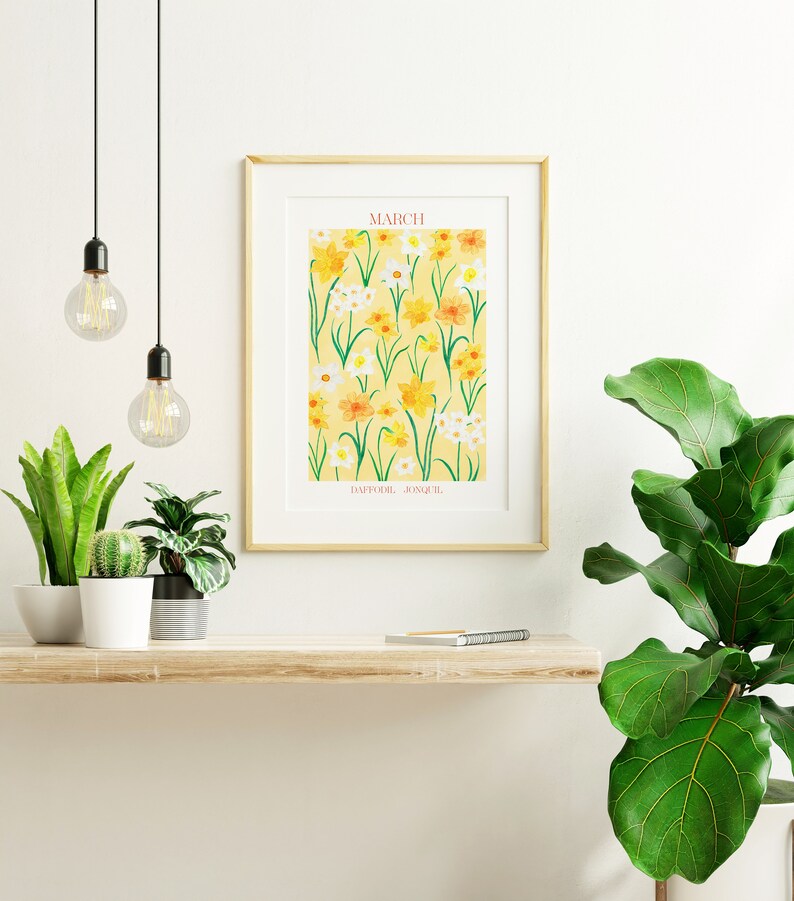 May include: A framed print with a yellow background and a pattern of yellow and white daffodils and jonquils. The print has the word "MARCH" and the words "DAFFODIL, JONQUIL" at the bottom. The print is hanging on a white wall above a wooden shelf with plants and a notebook.
