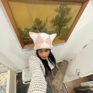 May include: A person wearing a pink and white crocheted hat with cat ears. The hat is made with a textured stitch and has a pink and white color scheme. The person is wearing a white sweater with a crocheted pattern.