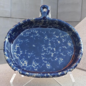 May include: A blue and white ceramic pie plate with a handle. The plate has a speckled glaze with a blue background and white speckles.