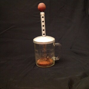 Pode incluir: Um copo de medição vintage de vidro com uma tampa de metal e uma pega de madeira no topo. O copo tem uma pega e marcas para medir em chávenas e onças. O copo está cheio de um pó castanho.