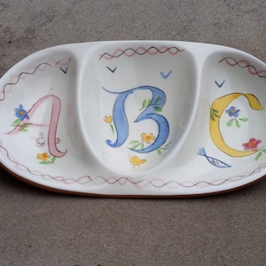 May include: A white ceramic serving dish with three sections, each decorated with a different letter of the alphabet: A, B, and C. The letters are painted in pink, blue, and yellow, respectively, and are surrounded by floral designs.