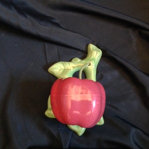 May include: A ceramic apple-shaped wall decoration. The apple is a vibrant red with a glossy finish, and it's attached to a green stem and leaves. The design is likely for kitchen or dining room decor.
