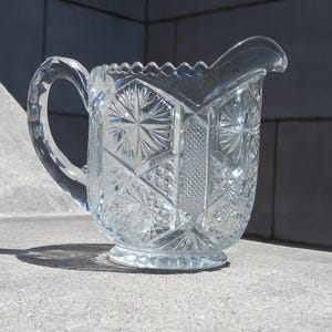 May include: Clear glass pitcher with a decorative, geometric pattern. The pitcher features a handle and a spout. The design includes starburst and diamond shapes. The pitcher is sitting on a concrete surface.