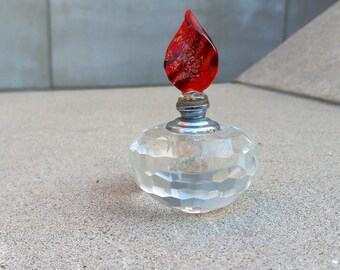May include: A clear, faceted glass perfume bottle with a red flame-shaped stopper. The stopper has a textured, marbled appearance with red and gold hues. The bottle has a round shape and a silver-colored metal base.