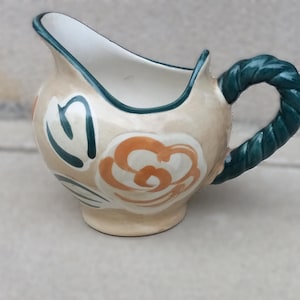Könnte beinhalten: Ein cremefarbener Keramik-Milchkännchen mit einem grünen, seilartigen Griff und einem floralen Design in Orange- und Grüntönen.