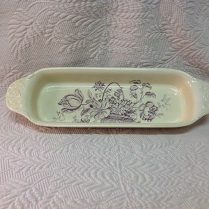 May include: A white ceramic serving dish with a purple floral design. The dish has two handles on the sides.