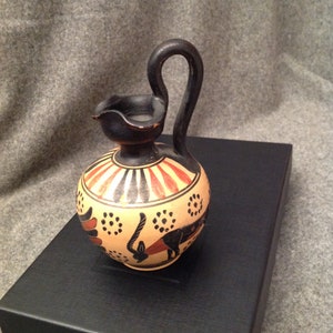 May include: A small, antique ceramic jug with a black handle and spout. The jug features a cream-colored body with red and black painted designs, including geometric patterns and stylized figures. The jug is displayed on a black surface.