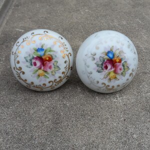 May include: Two white porcelain cabinet knobs with floral designs. Each knob features a central bouquet of pink, red, blue, and yellow flowers, surrounded by gold filigree. The knobs are round and have a glossy finish.