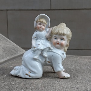 May include: A white porcelain figurine depicting a crawling figure with a smaller figure riding on its back. The figures have blonde hair, rosy cheeks, and are wearing white dresses. The figurine has gold accents and is set against a neutral background.
