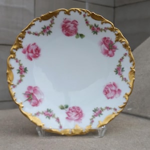 以下が含まれることがあります： 金色の縁取りとピンクのバラの花柄が施された白い磁器のプレート。プレートは波打った縁取りになっており、デザートや軽食を盛り付けるのに最適です。
