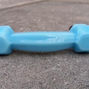May include: Two light blue dumbbells with a glossy finish are positioned on a textured gray surface. The dumbbells have a classic design with hexagonal ends and a curved handle. Fitness equipment.
