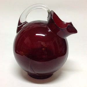 Puede incluir: Una jarra redonda de vidrio rojo rubí con un asa de vidrio transparente. La jarra tiene un cuerpo bulboso y un pico acampanado. El asa se arquea elegantemente desde la parte superior de la jarra hacia el lado. La jarra se utiliza para servir bebidas.