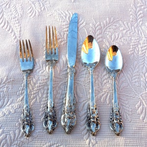 May include: A set of five antique silver-plated flatware pieces, including two forks, a knife, and two spoons. The silverware features ornate floral detailing on the handles and is arranged on a textured, light pink surface.