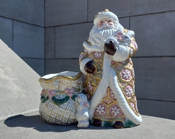 May include: A ceramic Santa Claus figurine with a white beard and a red and gold patterned coat. He holds a white bunny and stands next to a matching gift bag. The bag and Santa's coat have pink and gold accents.