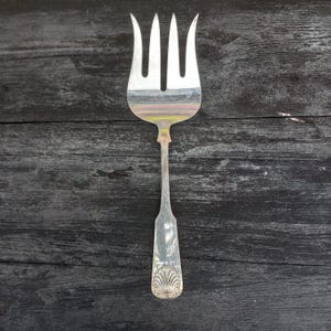 Op de afbeelding: Een zilverkleurige metalen serveervork met vier brede, gebogen tanden en een plat, taps toelopend handvat. De onderkant van het handvat is versierd met een verhoogd, waaiervormig schelpmotief. Het gepolijste oppervlak reflecteert het licht met subtiele kleuren.