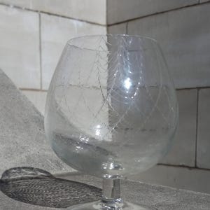 Könnte beinhalten: Ein klares Glasgefäß mit einem zarten, netzartigen Muster, das in die Oberfläche geätzt ist. Das Gefäß hat eine runde Schale und einen kurzen Stiel.