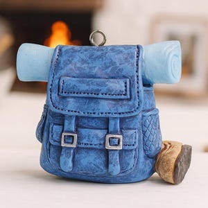 May include: A blue backpack ornament with a rolled-up sleeping bag attached. The backpack has silver buckles and a textured surface. A small brown boot is next to the backpack. The ornament has a silver loop for hanging.
