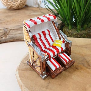 Könnte beinhalten: Ein Miniatur-Strandkorb mit rot-weiß gestreiften Kissen und Verdeck. Der Stuhl hat eine gewebte Korbrückenlehne und Seiten, mit Holz- und silberfarbenen Akzenten. Ein kleines gelb-weiß gestreiftes Kissen liegt auf dem Sitz.