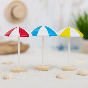 Könnte beinhalten: Drei Miniatur-Strandschirme mit weißen Stangen und Holzsockeln. Die Schirmspitzen sind rot und weiß, blau und weiß sowie gelb und weiß. Die Szene umfasst Muscheln und einen verschwommenen Hintergrund.