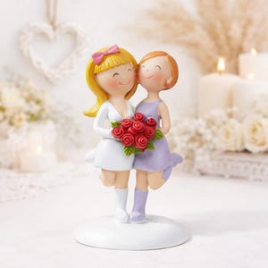 May include: A wedding cake topper featuring two smiling figures, one with blonde hair and a pink bow, the other with auburn hair, embracing. They hold a bouquet of red roses. The figures are dressed in a white dress and a lavender dress.