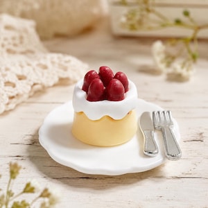 May include: A miniature round cake with a pale yellow base, topped with white frosting and five dark red berries. The cake sits on a small white scalloped plate alongside a tiny silver-colored metal knife and a matching fork on the right side.