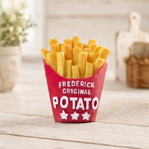May include: A red container filled with yellow french fries. The container features the white text FREDERICK ORIGINAL POTATO and three white stars at the bottom. The object is a decorative item with a textured finish, showcasing a classic fast food design.