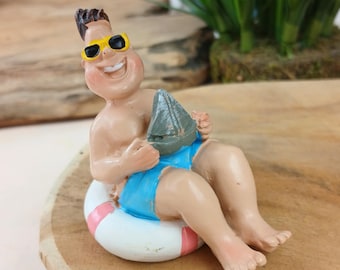May include: A figurine of a person in a blue swimsuit and yellow sunglasses, sitting in a white and pink inflatable ring. The person is holding a small gray sailboat. The figurine is on a wooden surface.